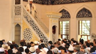 إمام يلقي خطبة بمسجد مزدحم بالمصلين والعمارة والزخارف الإسلامية بارزة.