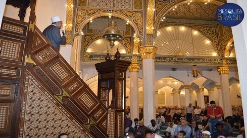 شخص يلقي خطبة على منبر مسجد مزخرف أمام مصلين جالسين.