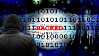 شخص بغطاء رأس يواجه شاشة تعرض كلمة "HACKED" وأرقام ثنائية، مفهوم الأمن السيبراني.