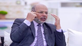 "An elderly man in a suit speaking and gesturing with his hands."