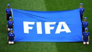 أشخاص يرتدون قبعات زرقاء يحملون لافتة "FIFA" على ملعب عشبي.