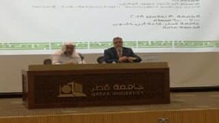 رجلان على منصة بجامعة قطر وشاشة خلفهما تعرض موضوع ندوة أكاديمية.