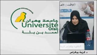 امرأة تتحدث في ميكروفون أمام لافتة جامعة وهران أحمد بن بلة.