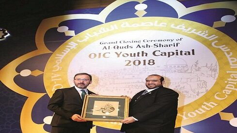 رجلان على منصة في حفل ختام عاصمة الشباب لمنظمة التعاون الإسلامي في القدس 2018 يحملان لوحة.