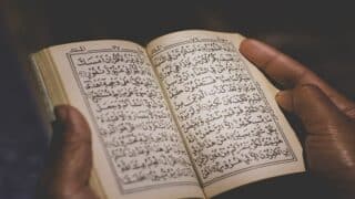 شخص يحمل كتاباً مفتوحاً يحتوي على نص ديني باللغة العربية بخط تقليدي.