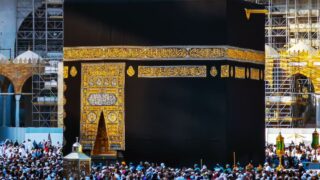 Kaaba with black cloth and gold calligraphy, surrounded by crowd and scaffolding.