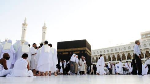 People in white gather by the Kaaba at Masjid al-Haram, Mecca; minarets in the background.