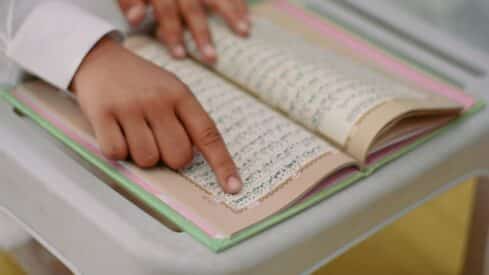 Person points at text in Quran book with intricate script on white surface, pink and green edges.