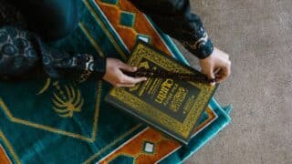 Person holds prayer beads over Quran on ornate green and orange prayer rug.