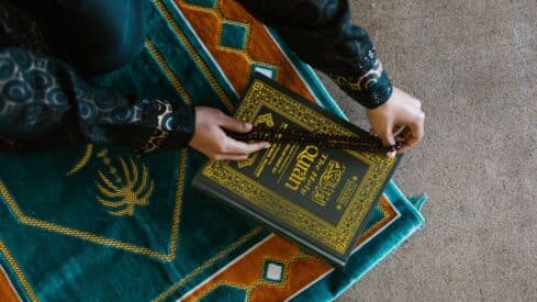 Person holds prayer beads over Quran on ornate green and orange prayer rug.