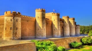 Historic stone castle with cylindrical towers, crenellated walls, flags under blue sky, greenery.