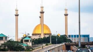 Large mosque with golden dome, four minarets, greenery, buildings, road, vehicles, cloudy sky.