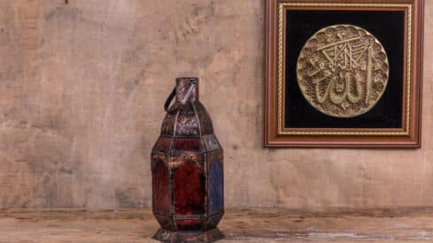 Decorative metal lantern with stained glass and framed Arabic calligraphy on wooden surface.