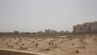 Large cemetery with unmarked graves and simple headstones, modern buildings in background.