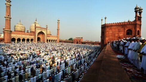 Large crowd in mosque courtyard praying, with arches and domes in the background.