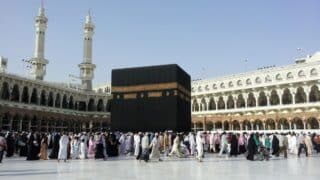 Crowd walking around the Kaaba in Masjid al-Haram courtyard, Mecca, with minarets visible.