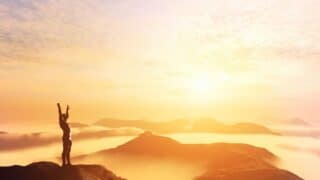 Person on mountain peak with arms raised, silhouetted against bright sunrise, misty hills in view.