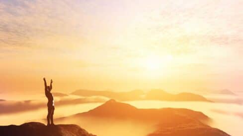 Person on mountain peak with arms raised, silhouetted against bright sunrise, misty hills in view.