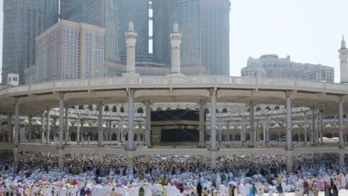 Large crowd around the Kaaba in Masjid al-Haram, Mecca, with minarets and tall buildings.