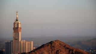 Tall clock-topped building with crescent spire behind rocky hill and hazy mountains.