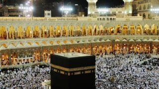 Aerial view of the Kaaba in Mecca, surrounded by a large crowd in a brightly lit mosque.