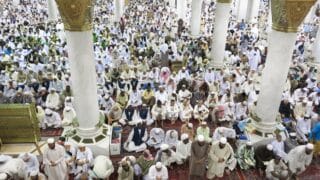 Large group gathered in a mosque, praying on carpeted floor, surrounded by tall columns.