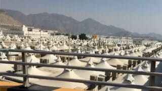 White tents arranged in rows in hajj mountains in background, railing in foreground, daytime setting.