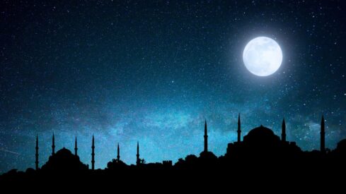 Silhouette of buildings with minarets under a starry night sky and a bright full moon