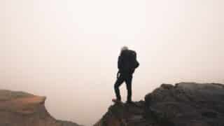 A person with a backpack standing on the edge of a rock in the middle of a thick fog.