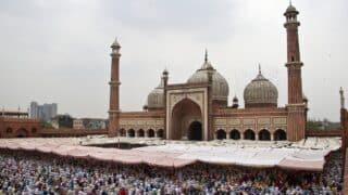 A large mosque with domes and minarets, gatherings under white umbrellas, a modern city in the background.