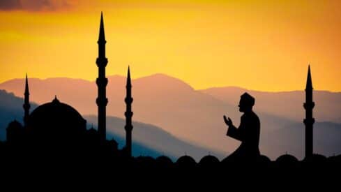 A picture of someone praying at sunset with the mountains and minarets of a mosque in the background.