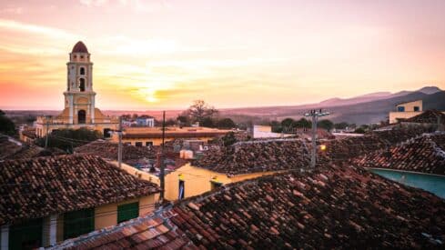 Sunset over an ancient city with a tile roof and a church tower, and a backdrop of mountains and trees.