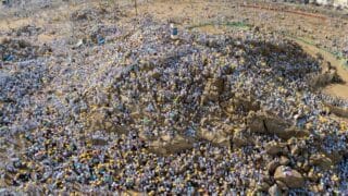 A large assembly dressed in white around a rocky hill during a religious event with colored umbrellas.