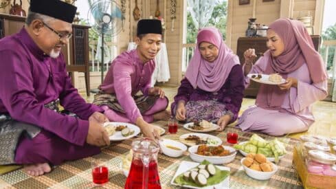 People in traditional clothes sit on the floor inside a house for a varied meal.