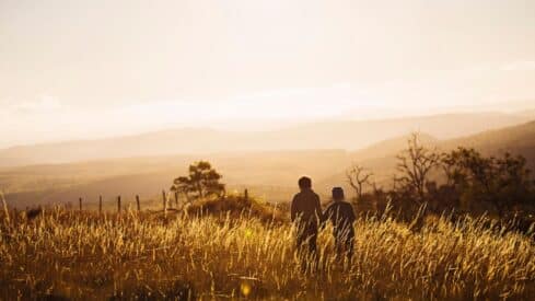 Two people walk in a sunset grass field with mountains and trees in the background.