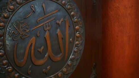 Round plaque with ornate Allah word in arabic calligraphy on a reddish-brown background.