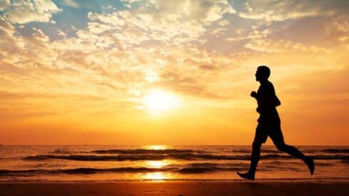 Person jogging along a beach at sunset, with waves and a vibrant, cloud-filled sky.