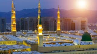 Mosque complex with illuminated minarets at sunset, surrounded by buildings and mountains.