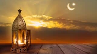 Decorative lantern with lit candle on wood, sunset sky with crescent moon and star.