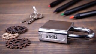 Silver padlock labeled "ETHICS" with gears, keys, and colored pens on wooden surface.