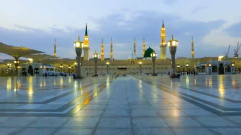 Open courtyard with marble tiles, lamp posts, minarets, green dome, cloudy sky, and umbrellas.