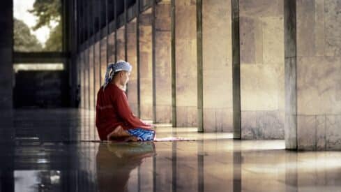 Person kneels and prays on a mat in a sunlit corridor with stone columns and glossy floor.