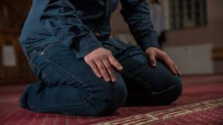 Person kneeling on patterned carpet, wearing dark jeans and long-sleeved shirt, hands on knees.