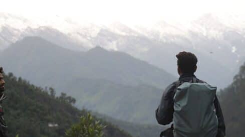 Person with a large backpack on a forested mountainside, snowy peaks visible in the distance.