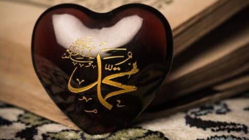 Dark heart-shaped object with gold Arabic calligraphy mohammed word; open book in background.