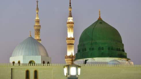 prophet mosque domes (green, white) and illuminated minaret against an evening sky.