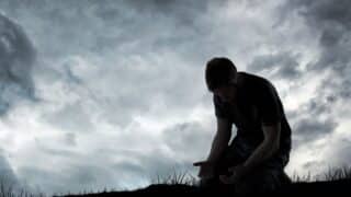 Silhouette of kneeling person with palms up against cloudy sky, grass visible in foreground.