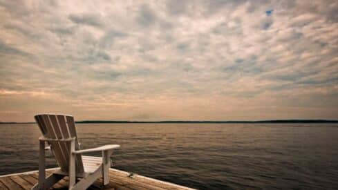 White Adirondack chair on dock overlooking calm water with cloudy sky and distant horizon.