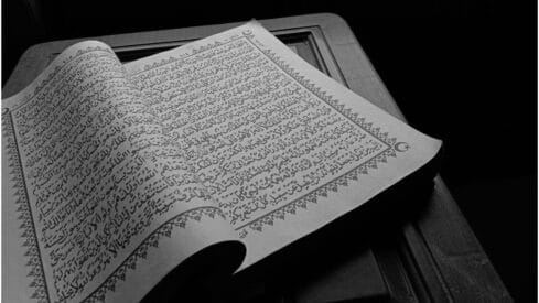 Open Quran page with Arabic text on wooden stand, ornate borders, black and white image.