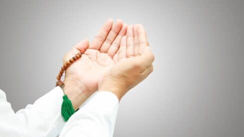 Hands in prayer pose with brown beads and green tassel on a light gray background.